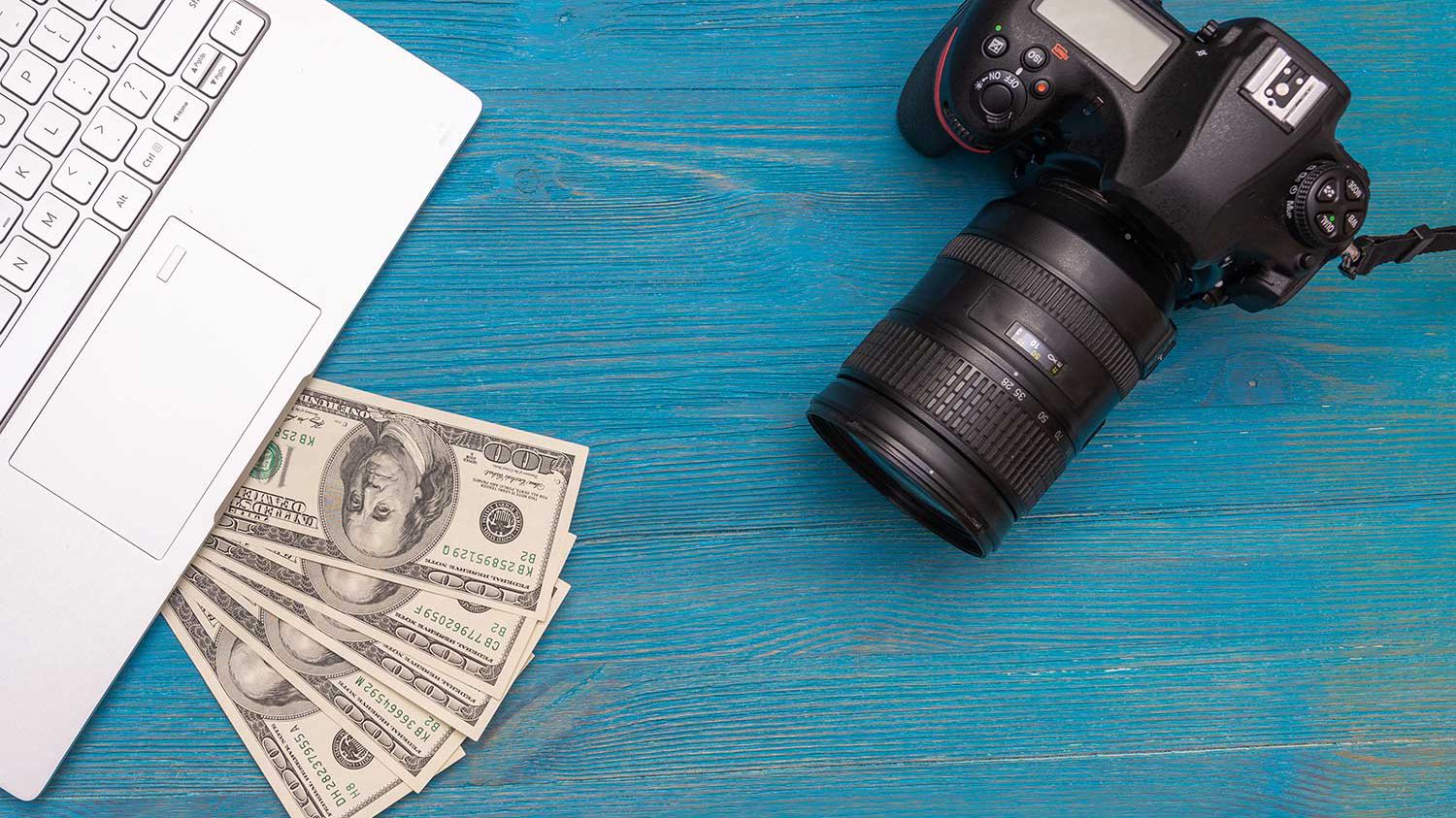 دوربین عکاسی به همراه دلار و لپ تاپ بر روی میز آبی
